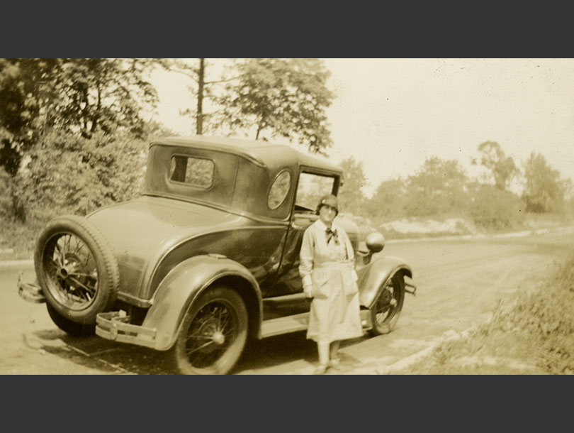 White female nurse in uniform standing beside a Ford car on a rural road.