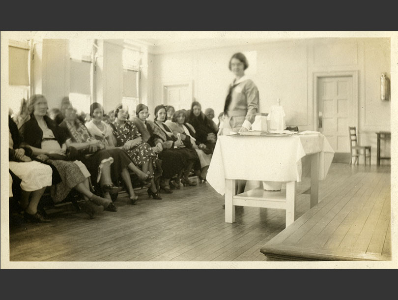 A uniformed, White, female nurse standing next to a table with an audience of seated women in front of her.