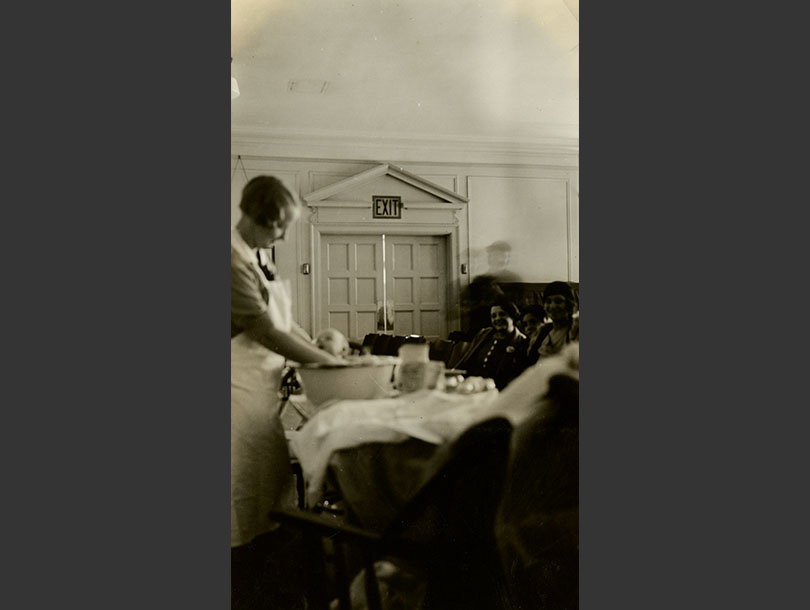 White female nurse bathing an infant in a basin while mothers observe.