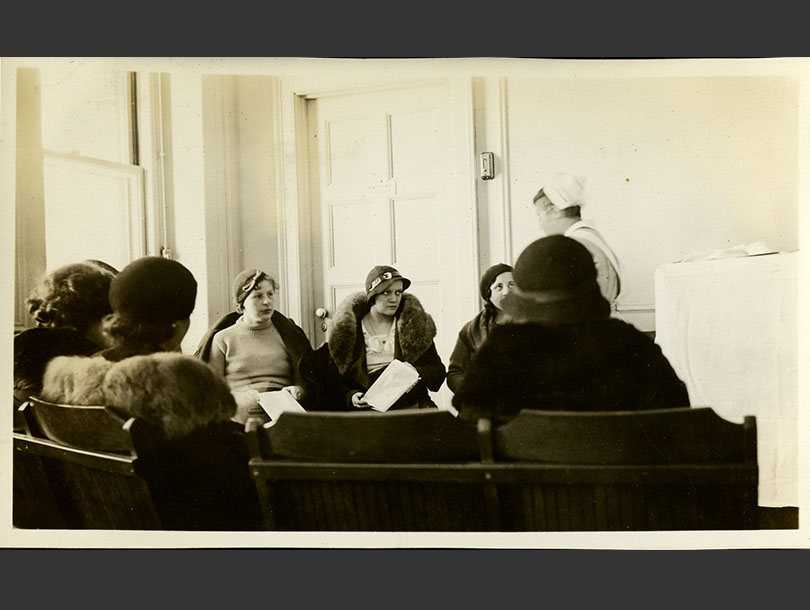 Sitting in a semi-circle, 7 females in coats watch a White female nurse in uniform conducting class.