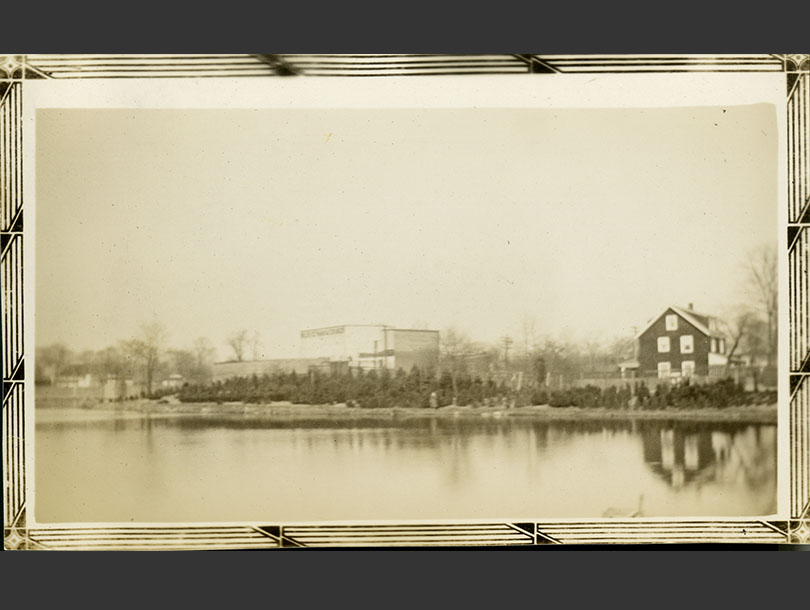 Pond with three-story shingled home and warehouse building in background.