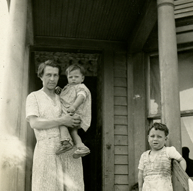 A mother, holding a toddler, and a young boy stand at the top step of wooden frame house.