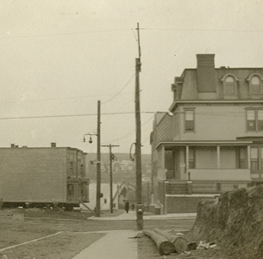 In the middle of a street a two-story house rests while being moved, two other houses nearby.