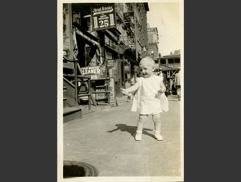 An 11-month White girl standing on a street in 1930s New York, signs for businesses in background.