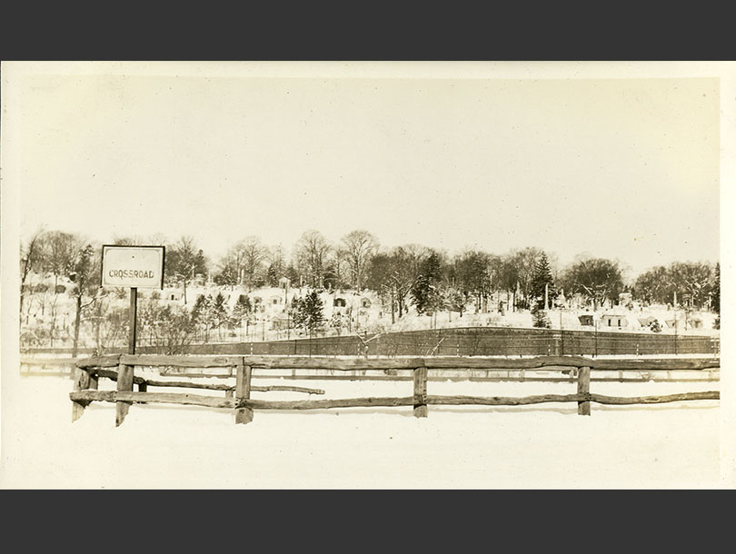 Snowy roadside with a split-rail fence and a cemetery up a low hill beyond.