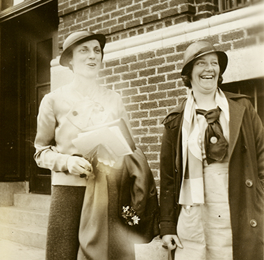 Two, White females in cloche hats standing near a brick building. Miss Giles is in uniform.