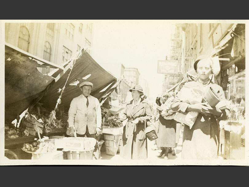 Female nurse in coat and cloche hat making a purchase from a man at a pushcart in Lower Manhattan.