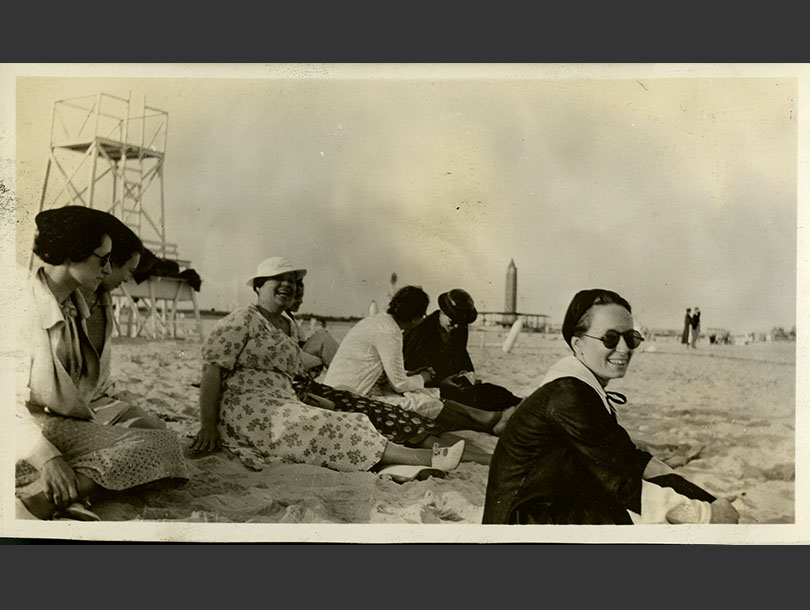 Seven women in dresses sitting on Jones Beach, life guard stand and obelisk water tower in background.