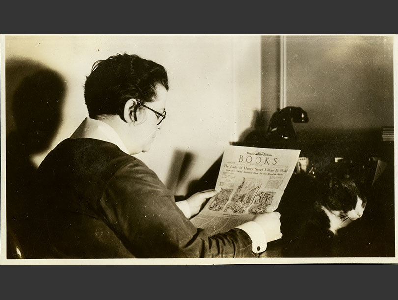 Profile view of a White, woman reading the <em>Herald Tribune<em> at a desk, nearby a cat and telephone.