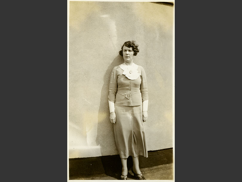 A White woman in a two-piece dress with white shawl collar standing full-length against a wall.