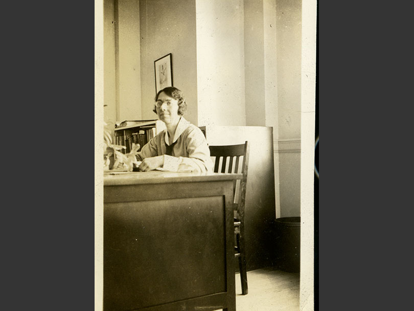 White, female nurse with glasses in a nursing uniform, sitting behind a desk in an office.